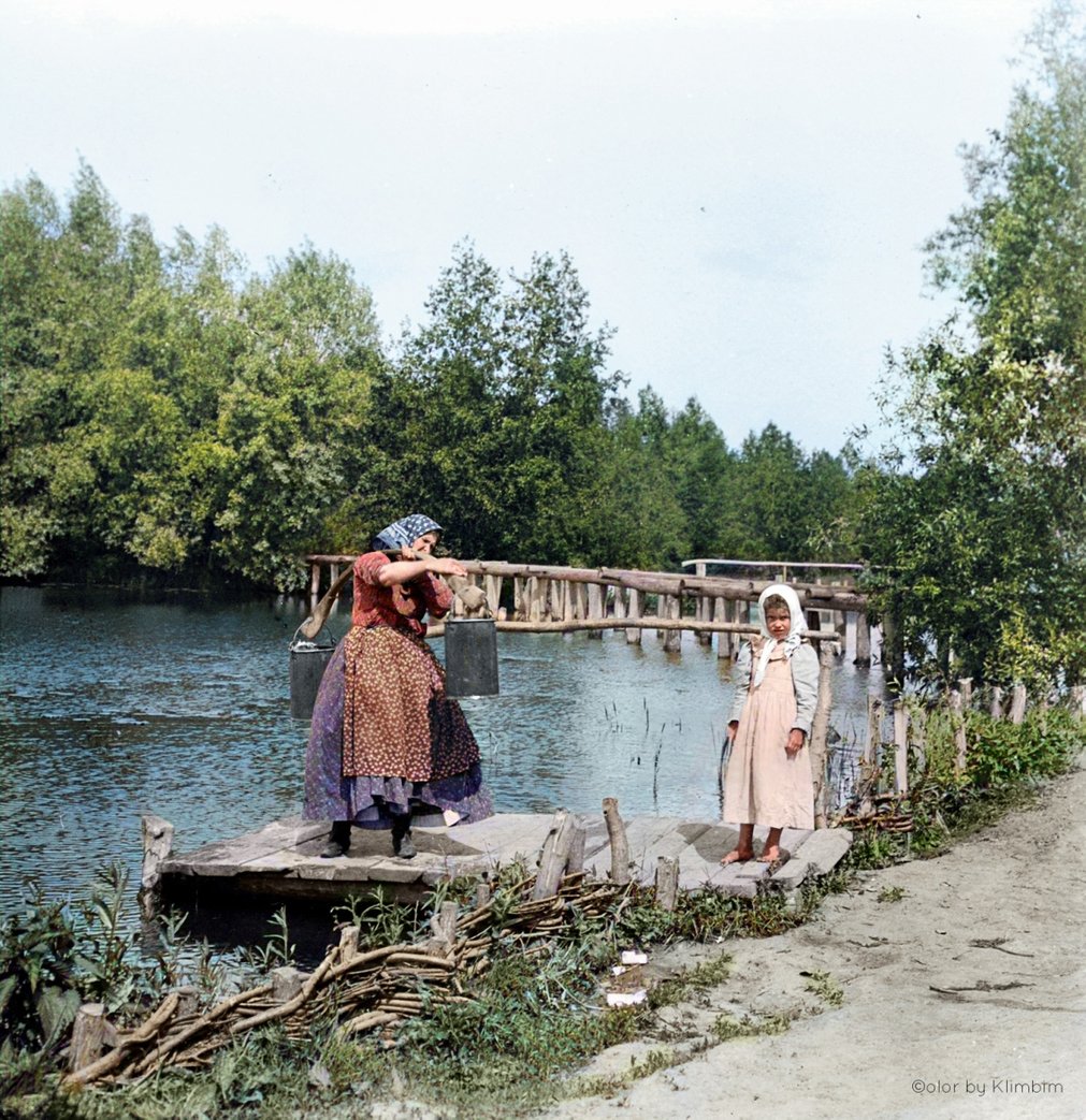 Peasant-woman-stands-on-wooden-jetty-next-to-river-carrying-two-buckets-of-water-on-yoke,-a-young-girl-also-stands-on-the-jetty,-Russia-1900s.jpg