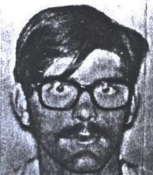 A grainy black-and-white photo of a young white man with straight though slightly unkempt hair, a mustache, and bespectacled eyes which stare straight ahead at the camera